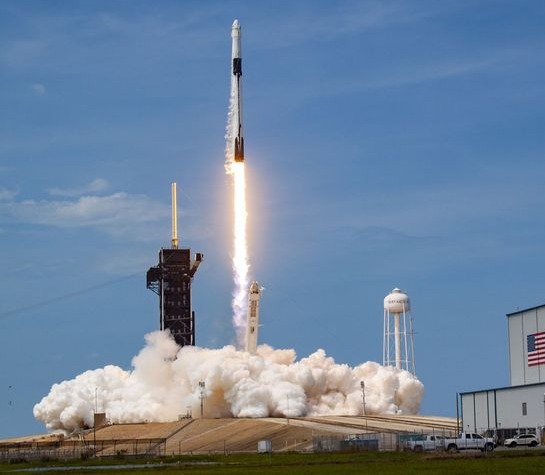 A launch vehicle lifting off from a coastal pad, exhaust plume spreading across the ground below.